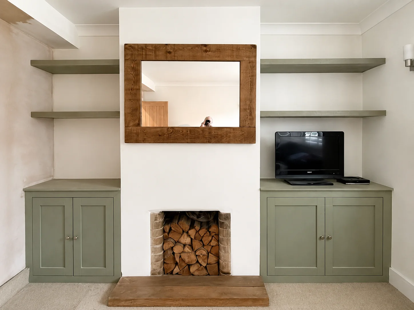 Green shaker alcove cupboards and floating shelves either side of a fireplace with oak hearth